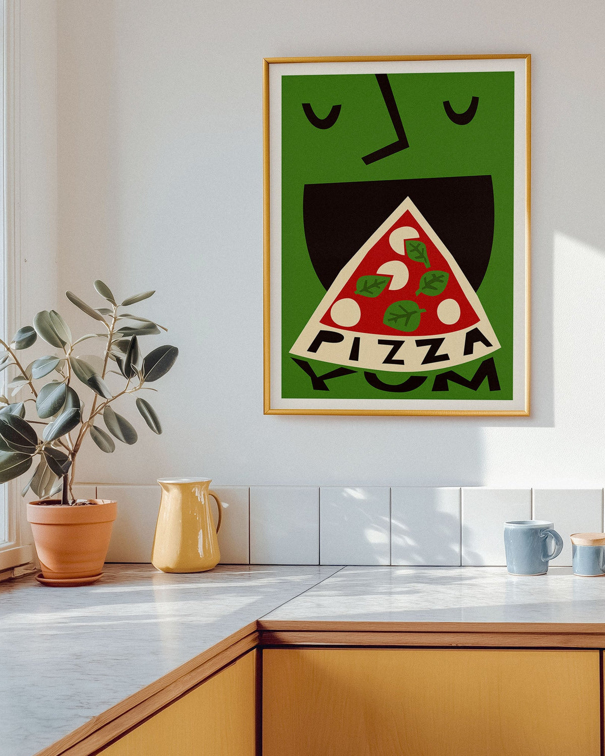 A framed art print featuring a stylized green face eating a slice of pizza with the word 'PIZZA' inside, hanging on a white kitchen wall above a counter with a plant and mugs.