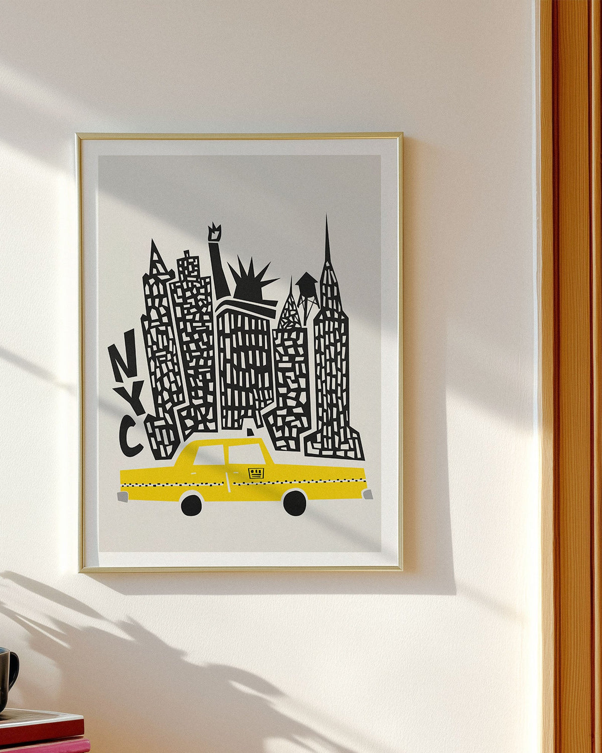 A framed art print featuring a stylized New York skyline with a yellow taxi and black architectural details on a light gray background, hung on a cream-colored wall next to a window.