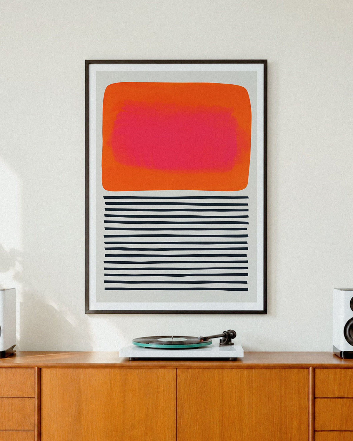 A framed abstract art print 'Sunset Ripples' featuring a bright orange and pink rectangle above black horizontal stripes, displayed on a wooden cabinet in a modern room setting with speakers and a record player.