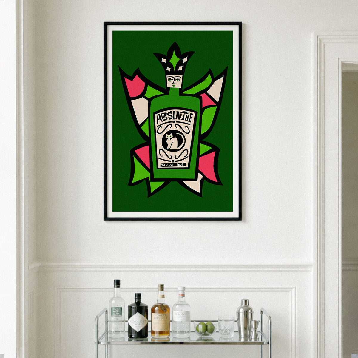 A framed wall art print featuring a stylized green Absinthe bottle illustration with pink, cream, and black graphic elements, hung on a white paneled wall above a glass bar cart with bottles and glasses.