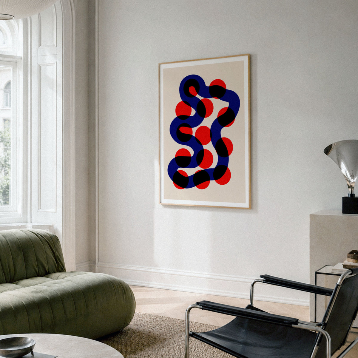 A framed abstract geometric art print with colorful twisting shape hangs on a light gray wall in a modern living room setting with a green sofa and black chair.