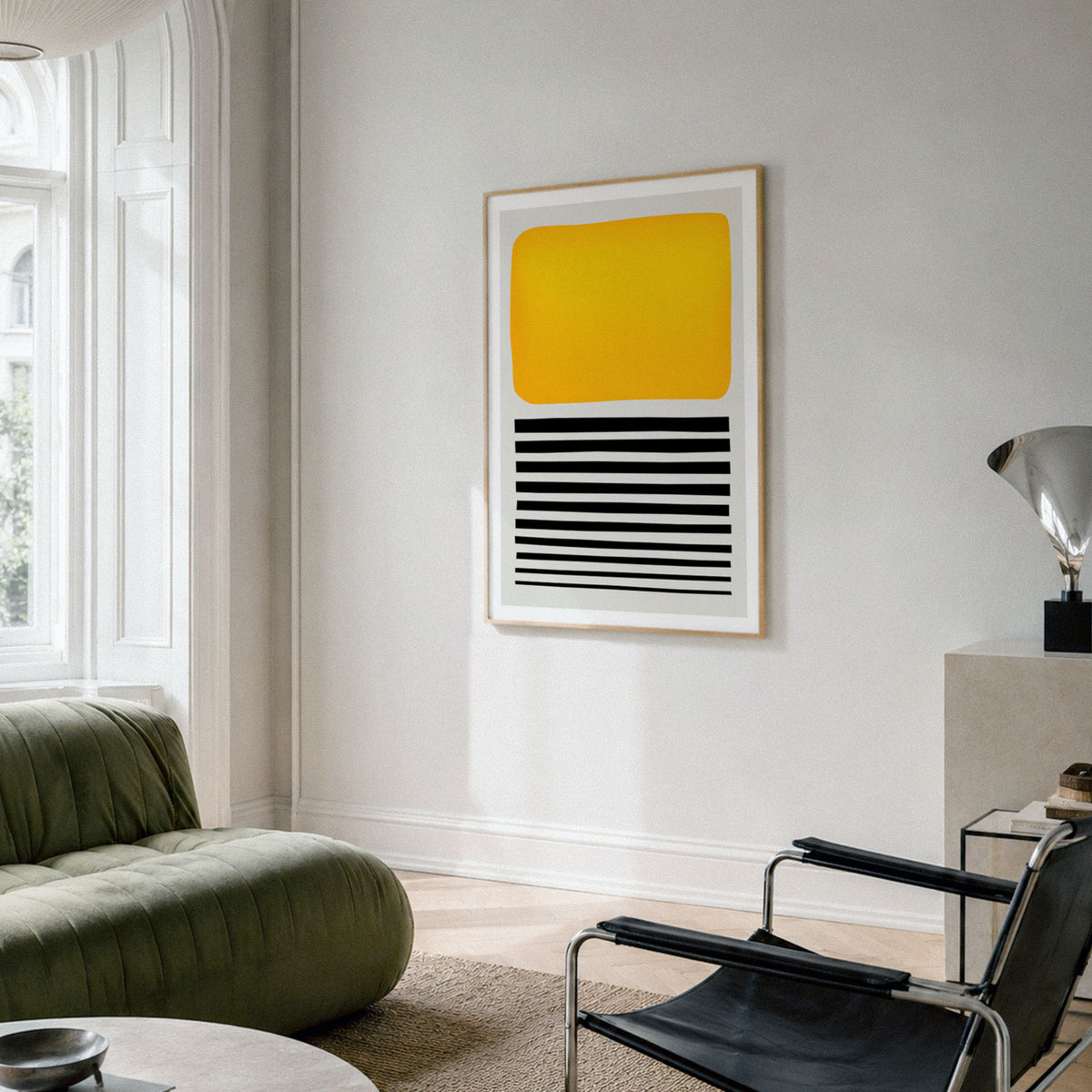 A framed abstract wall art print hangs on a light gray wall in a modern living room. The artwork features a large yellow rounded rectangle on the upper half and horizontal black stripes on the lower half, set in a thin light wood frame.