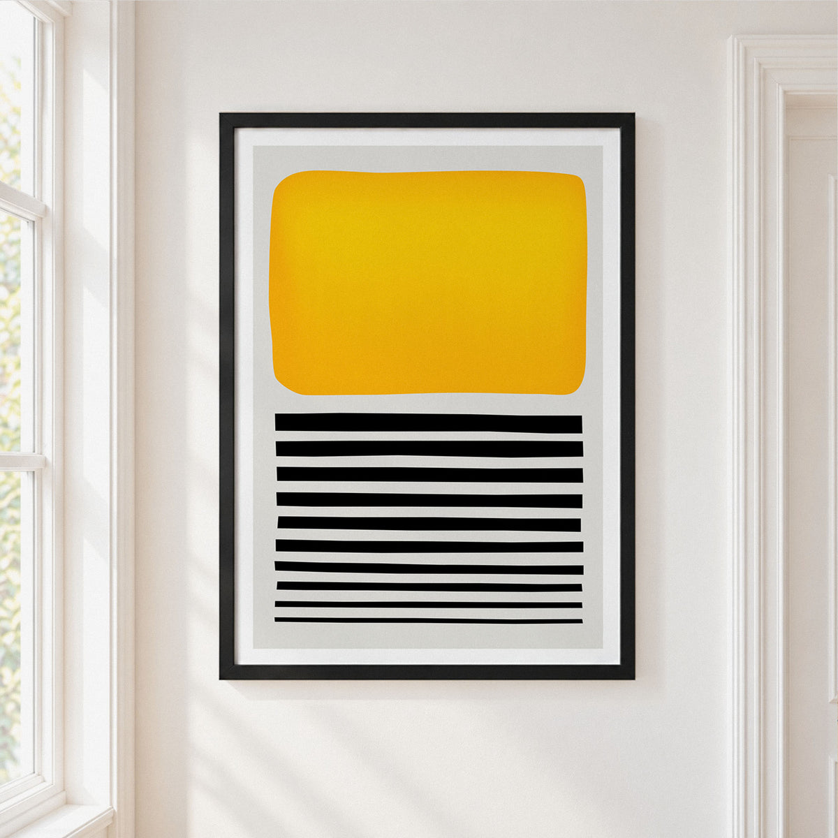 A framed abstract art print featuring a large yellow rounded rectangle above black horizontal stripes on a white background, hung on a white wall next to a window.