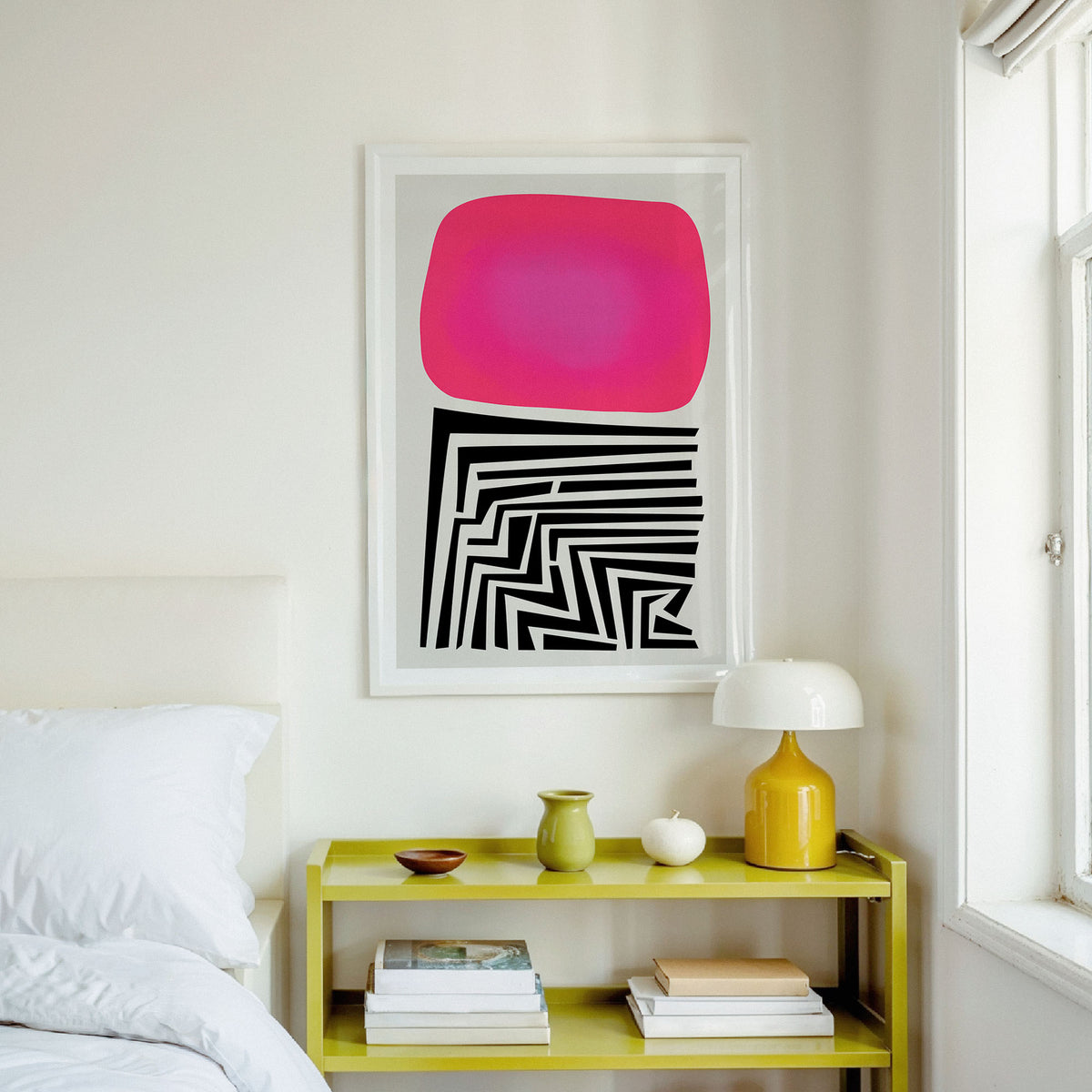 A framed abstract art print featuring a bright pink shape above black geometric lines, displayed on a white wall above a lime green nightstand in a bedroom setting.