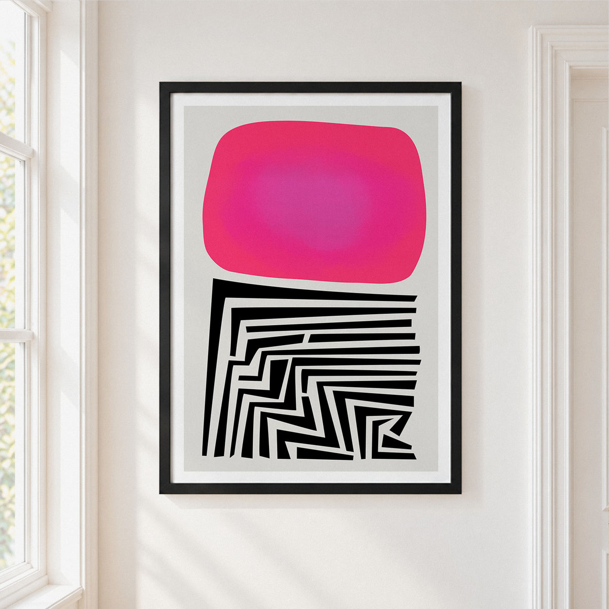 A framed abstract art print hanging on a white wall next to a window. The print features a bright pink rounded rectangle on a light gray background, with a black and white geometric maze-like pattern below it, enclosed in a black frame.