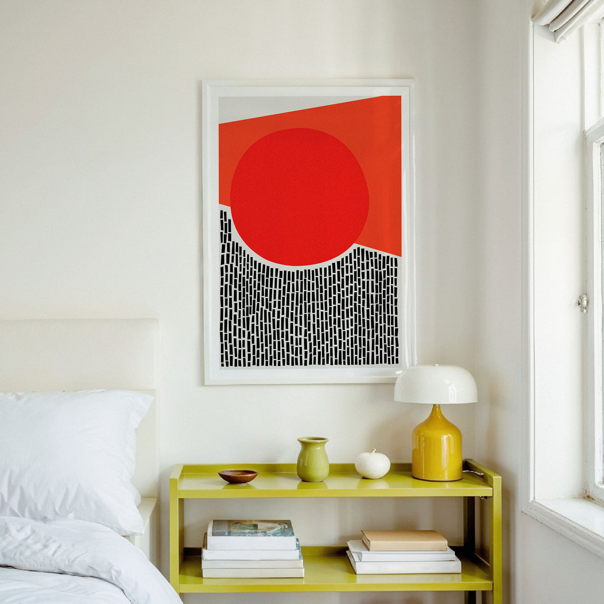 A framed abstract art print with a bold red circle and geometric shapes hangs on a white wall above a lime green nightstand in a bright bedroom setting.