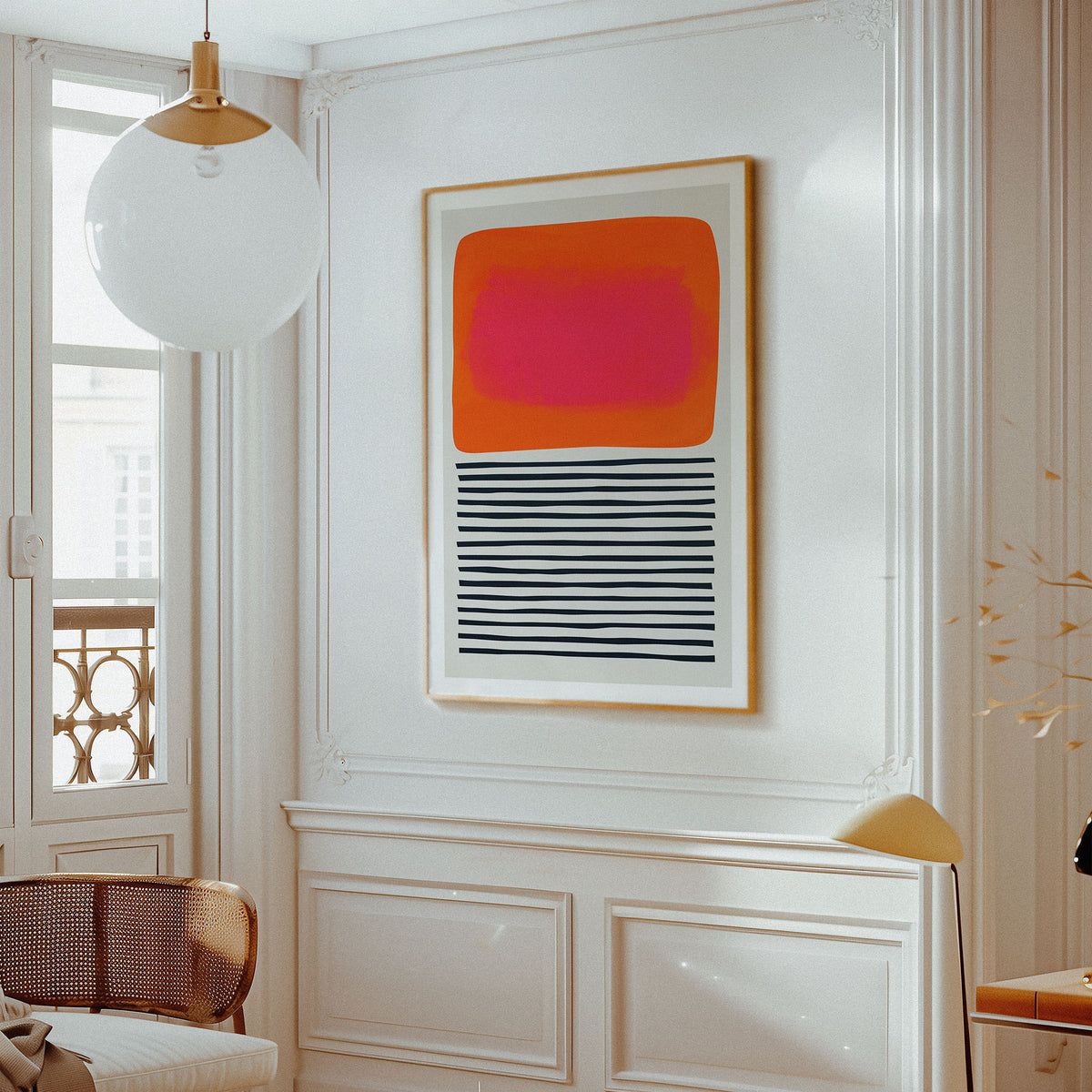 A framed abstract art print hangs on a white paneled wall in a bright, elegant room. The artwork features a vibrant pink and orange block on the top half and black horizontal stripes on the bottom half, set in a light wood frame.