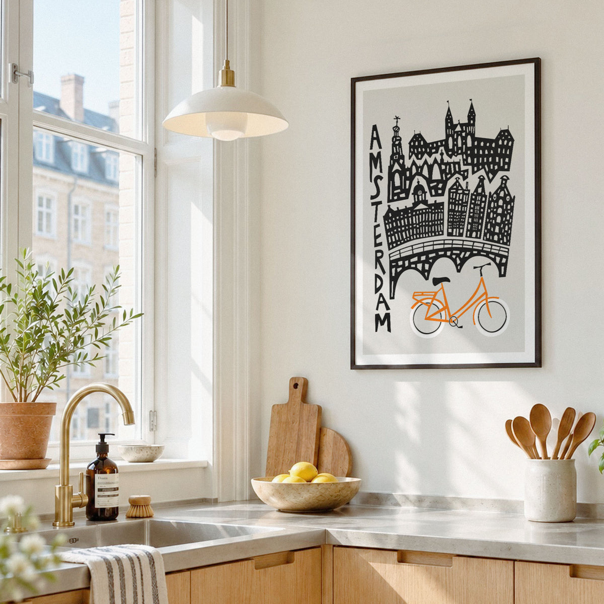 A framed pop-art style poster featuring a bicycle and black buildings, hanging on a white kitchen wall above a countertop with a sink, plants, and utensils.