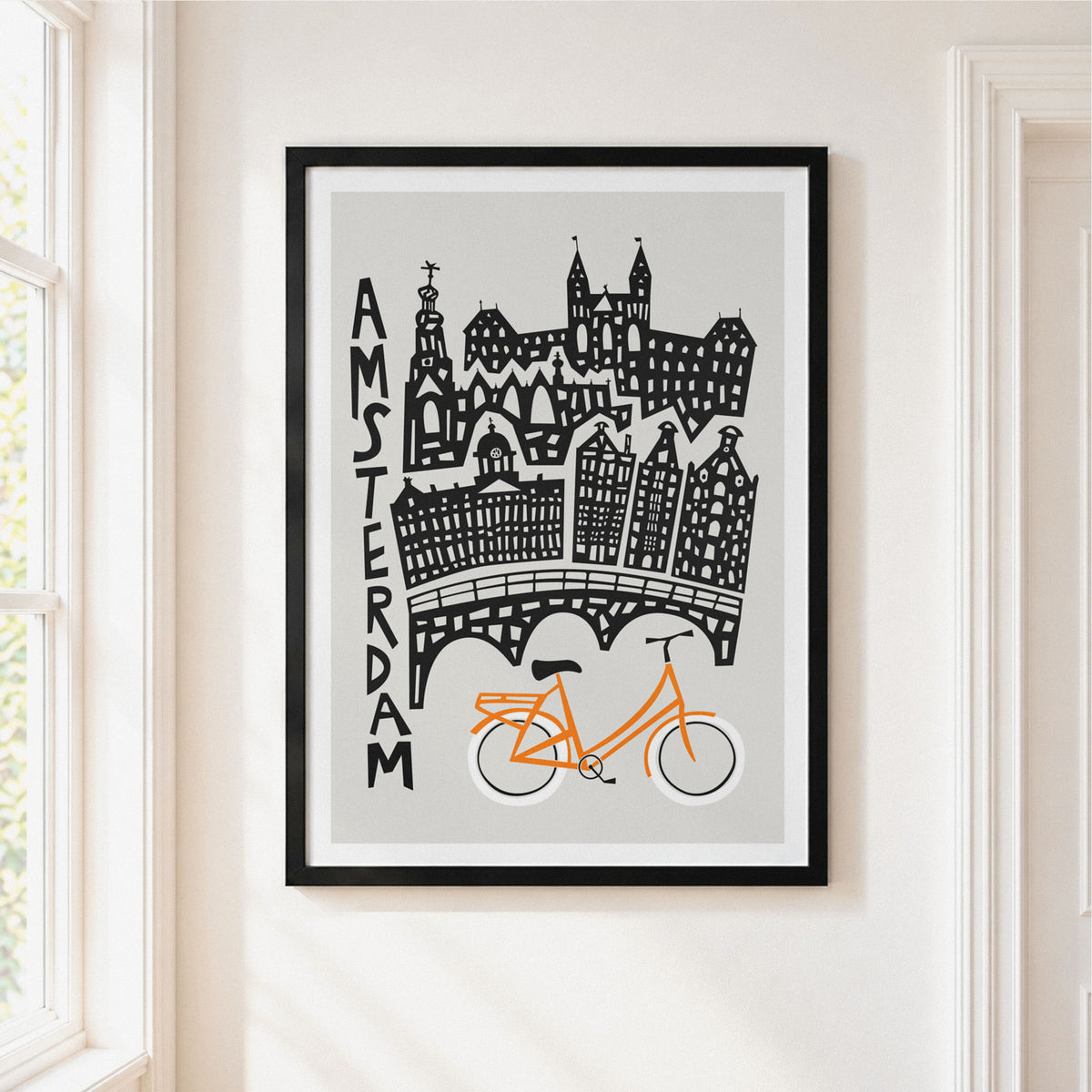 A framed art print featuring a stylized Amsterdam skyline with an orange bike and black architectural details on a light gray background, hung on a cream-colored wall next to a window.