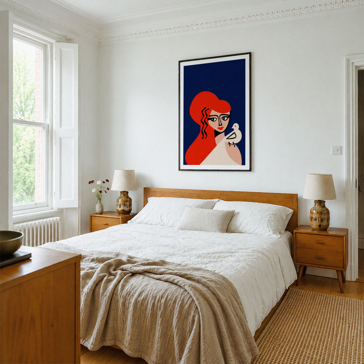A framed art print featuring a stylized illustration of Aphrodite with red hair against a dark blue background, displayed on a white wall above a wooden bed in a bright, modern bedroom.