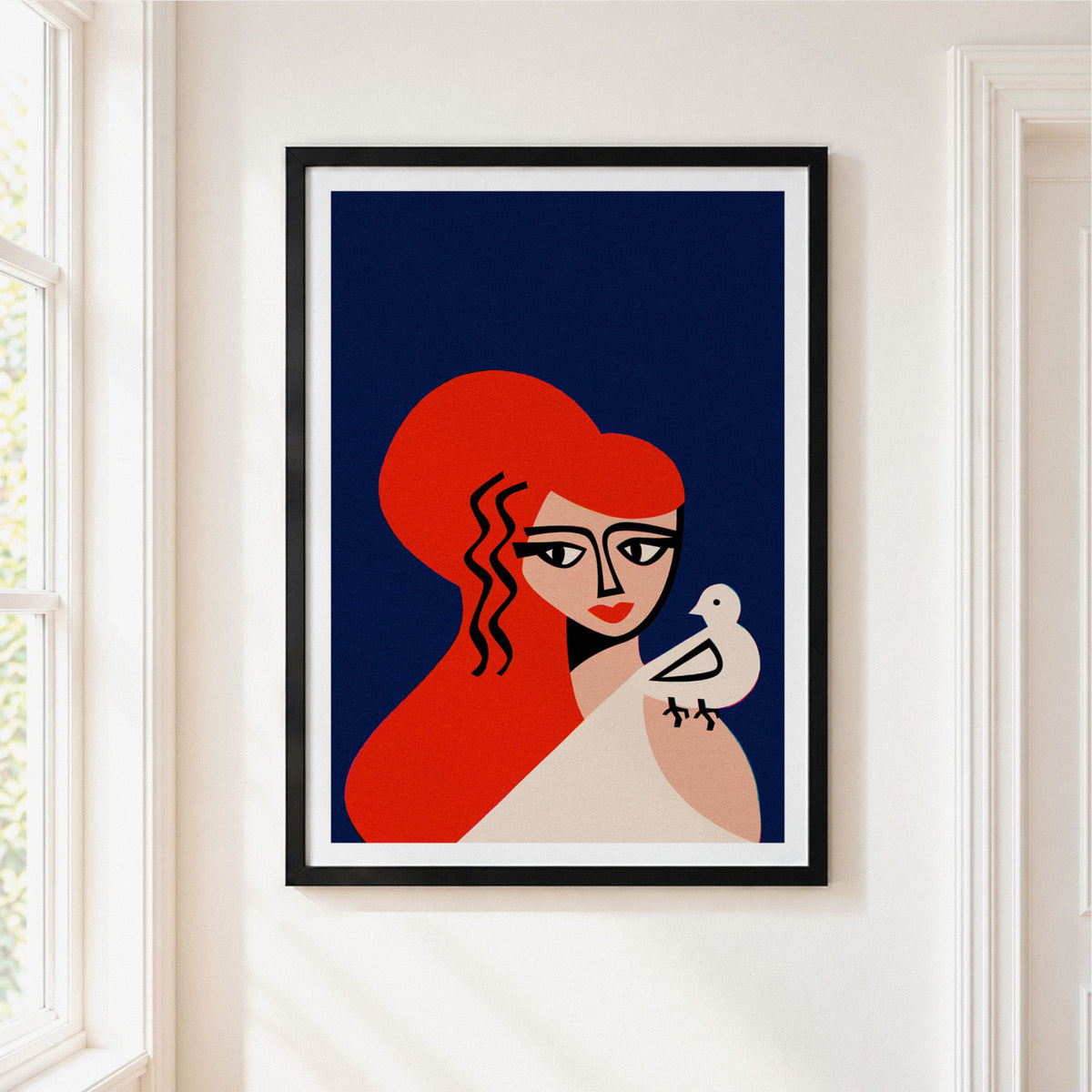 A framed art print featuring a stylized illustration of Aphrodite with vibrant red hair and a white bird, displayed on a clean white wall next to a window.