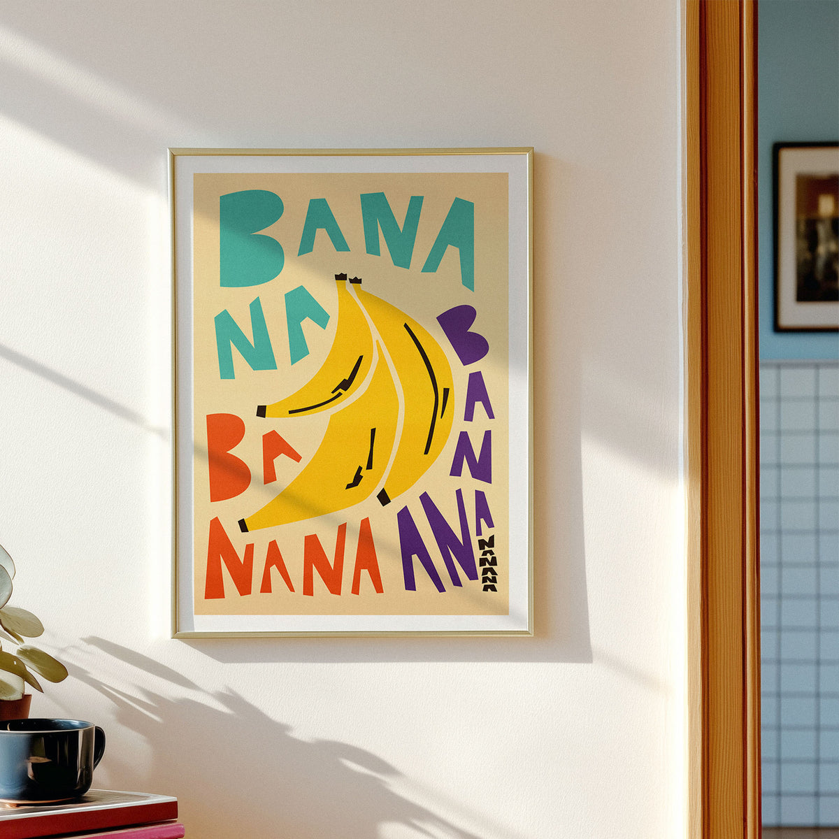 A framed art print featuring a retro-style banana illustration with the word 'BANANA' repeated in colorful lettering, hanging on a white wall in a sunlit room.
