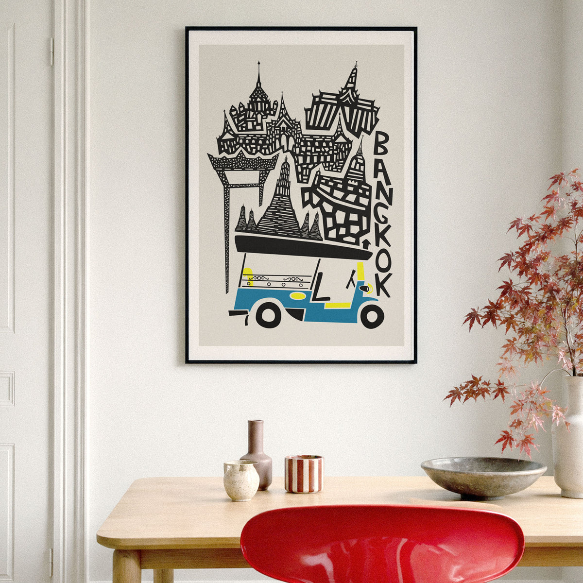 A framed travel poster of Bangkok hanging on a light gray wall above a wooden table with decorative vases, a bowl, and a red chair. The poster features a black and white illustration of Bangkok landmarks and a blue-and-yellow tuk-tuk, with the word 'BANGKOK' written vertically.