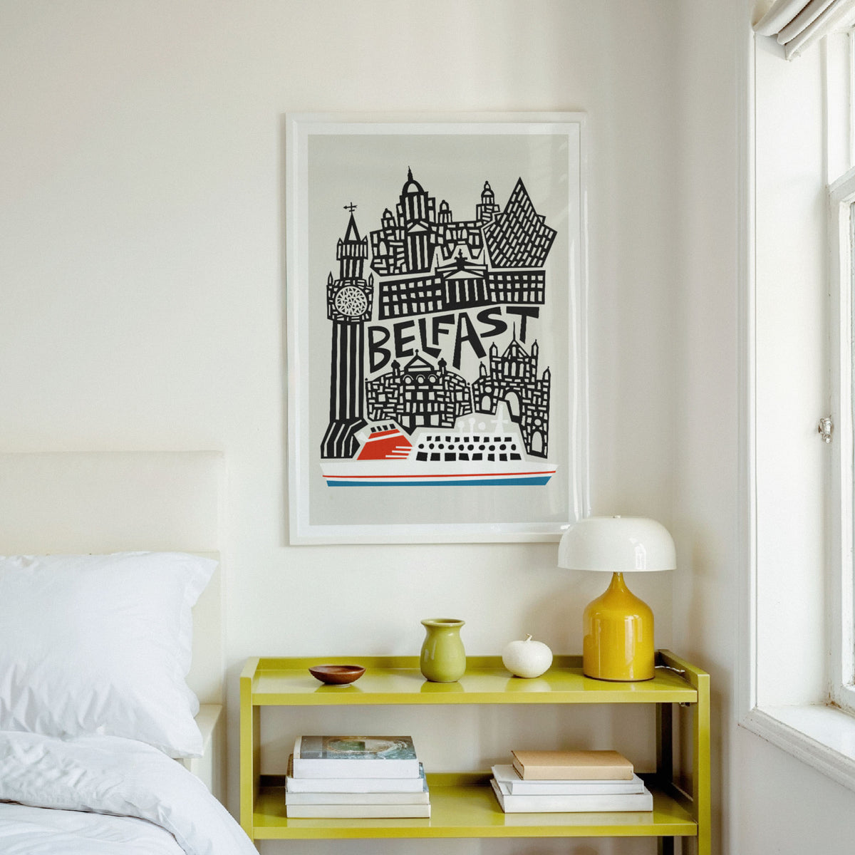A framed art print featuring a black and white illustration of Belfast's skyline with a red boat, hanging on a white wall above a yellow bedside table in a bright bedroom.