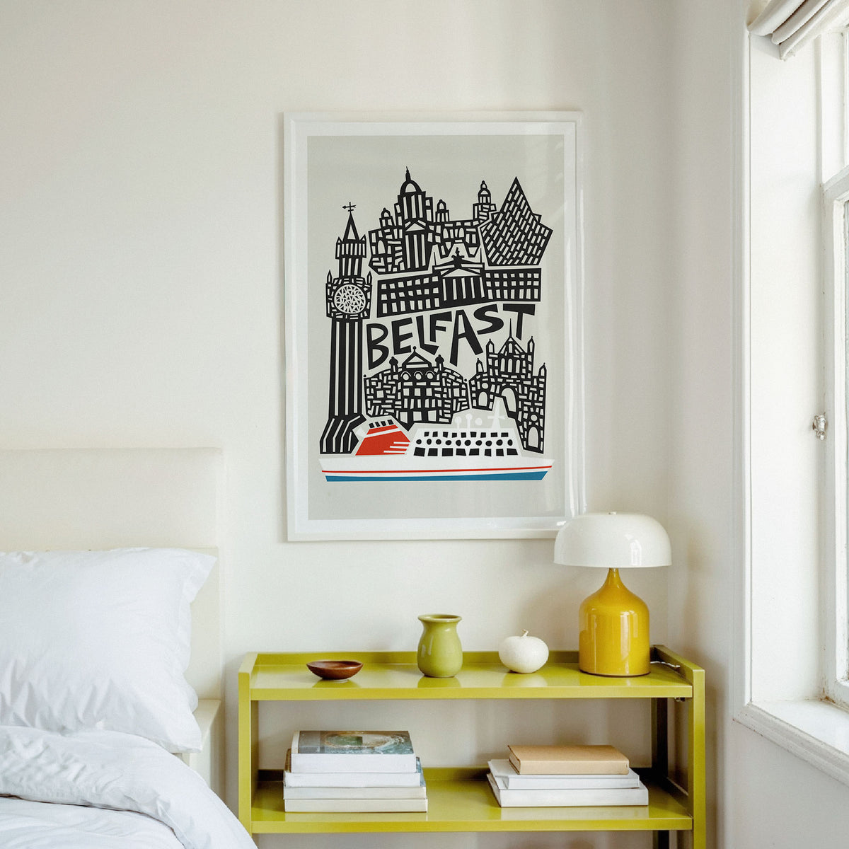 A framed art print featuring a black and white illustration of Belfast's skyline with a red boat, hanging on a white wall above a yellow bedside table in a bright bedroom.
