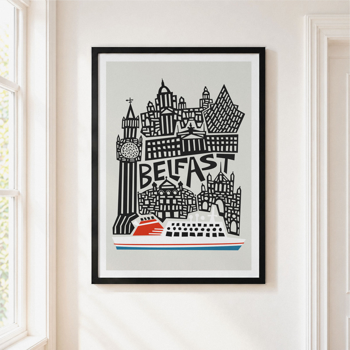 A framed art print featuring a stylized black and white illustration of Belfast landmarks with a red and white ferry at the bottom, hanging on a light beige wall next to a window.
