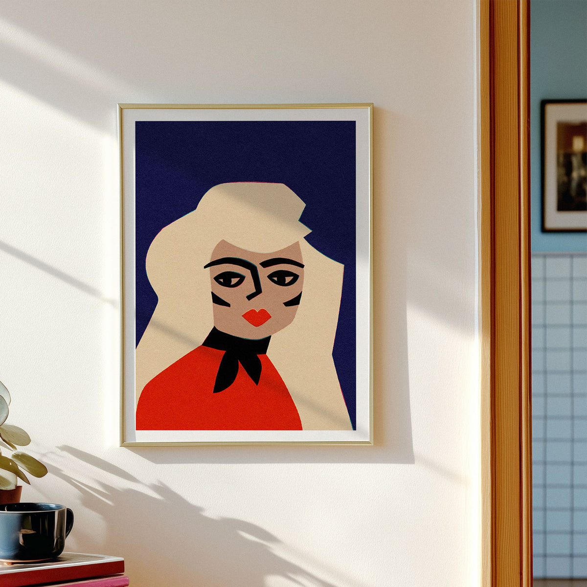 A framed art print featuring a stylized, geometric portrait of a woman in red and cream against a dark blue background, hanging on a white wall in a sunlit room.
