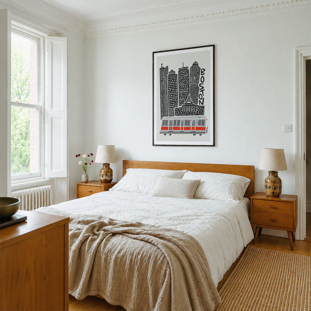 A styled bedroom interior featuring a wooden bed with white bedding and a beige linen throw, flanked by matching wooden nightstands with lamps. A framed black and white Boston cityscape print hangs on the white wall above the bed.