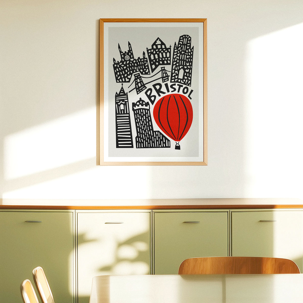 A framed art print featuring a stylized black and white illustration of Brighton landmarks with a bright blue scooter, hanging on a light beige wall above green cabinets.