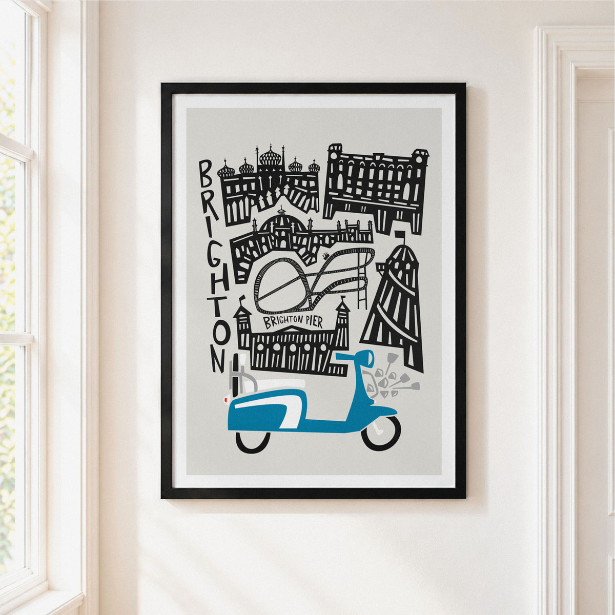 A framed art print featuring a stylized black and white illustration of Brighton landmarks with a blue scooter at the bottom, displayed on a cream-colored wall next to a window.