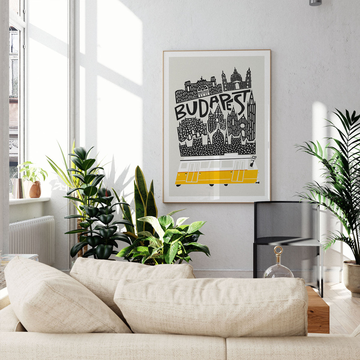 A framed Budapest cityscape art print hangs on a light gray wall above a beige sofa in a bright, modern living room with potted plants and natural sunlight.