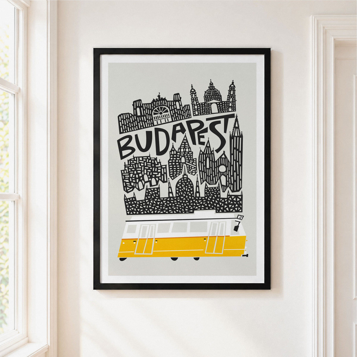 A framed Budapest travel art print hanging on a cream-colored wall next to a window. The poster features a black and white vector illustration of the Budapest skyline with the word 'BUDAPEST' written across it, and a yellow and white tram at the bottom.