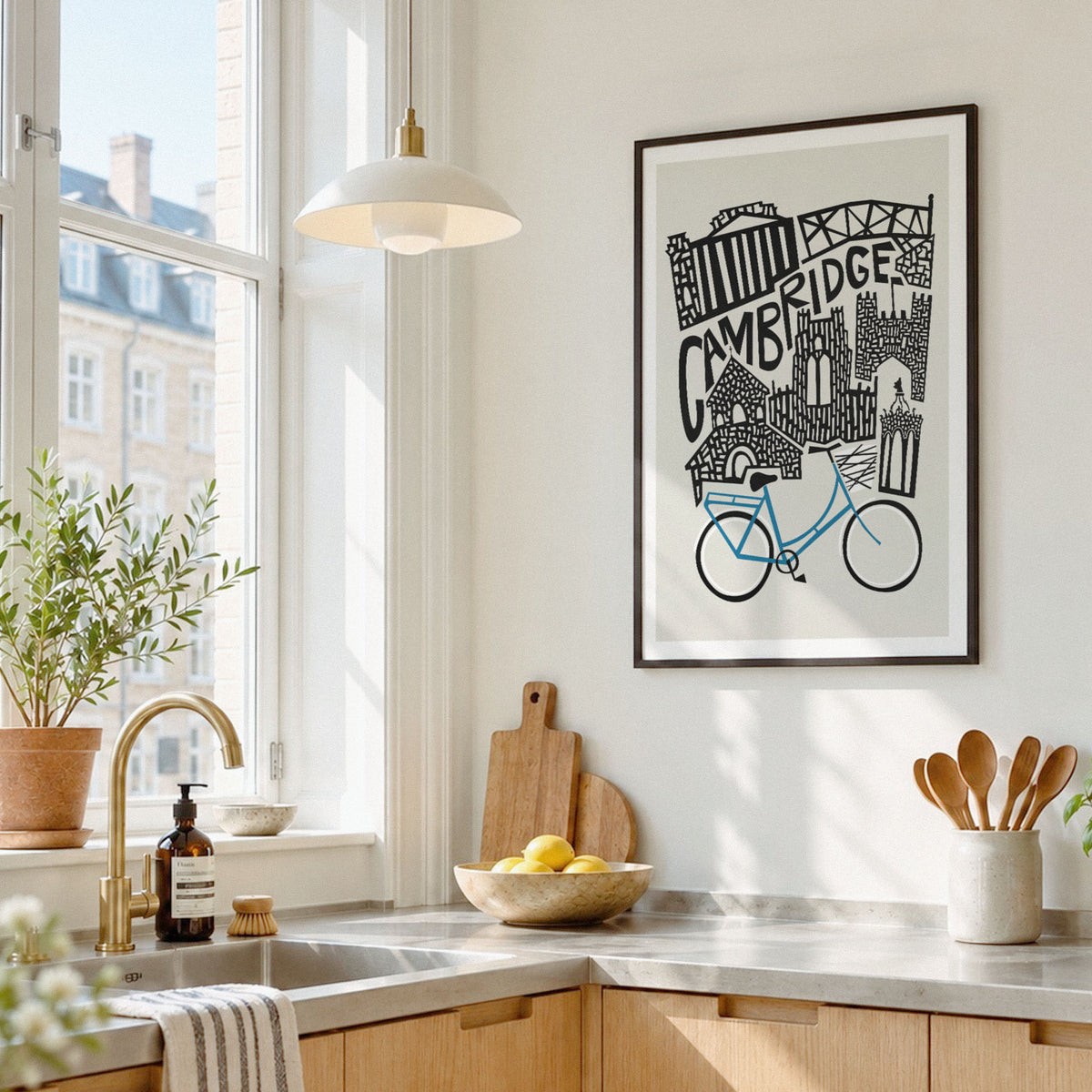 A framed black and white Cambridge cityscape art print with a blue bicycle illustration, hanging on a light wall in a bright, modern kitchen.