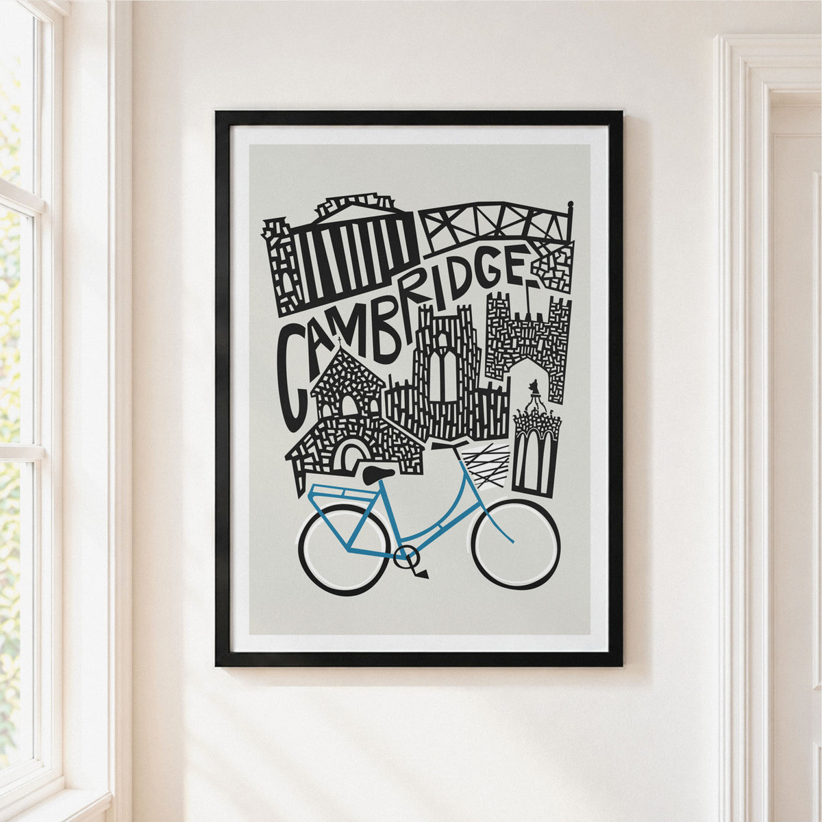 A framed art print featuring a black and white hand-drawn illustration of Cambridge landmarks with a blue bicycle, hanging on a warm off-white wall next to a window.