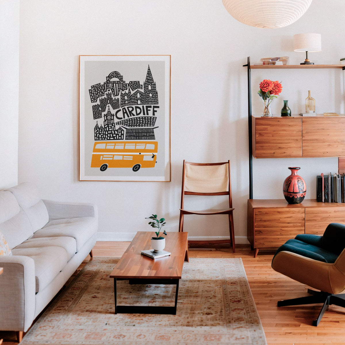 A framed art print featuring a black and white cityscape of Cardiff with a yellow bus, displayed on a white wall in a modern, sunlit living room with a beige sofa, wooden coffee table, and mid-century modern furniture.
