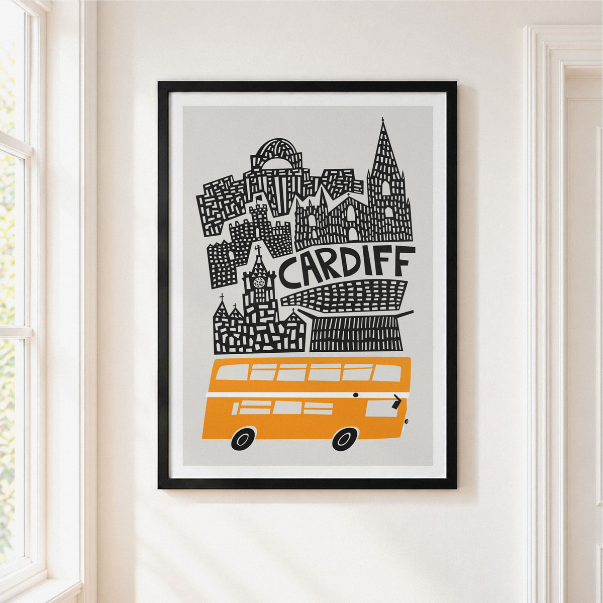 A framed art print featuring a stylized black and white Cardiff skyline with an orange double-decker bus, hanging on a light beige wall next to a window.
