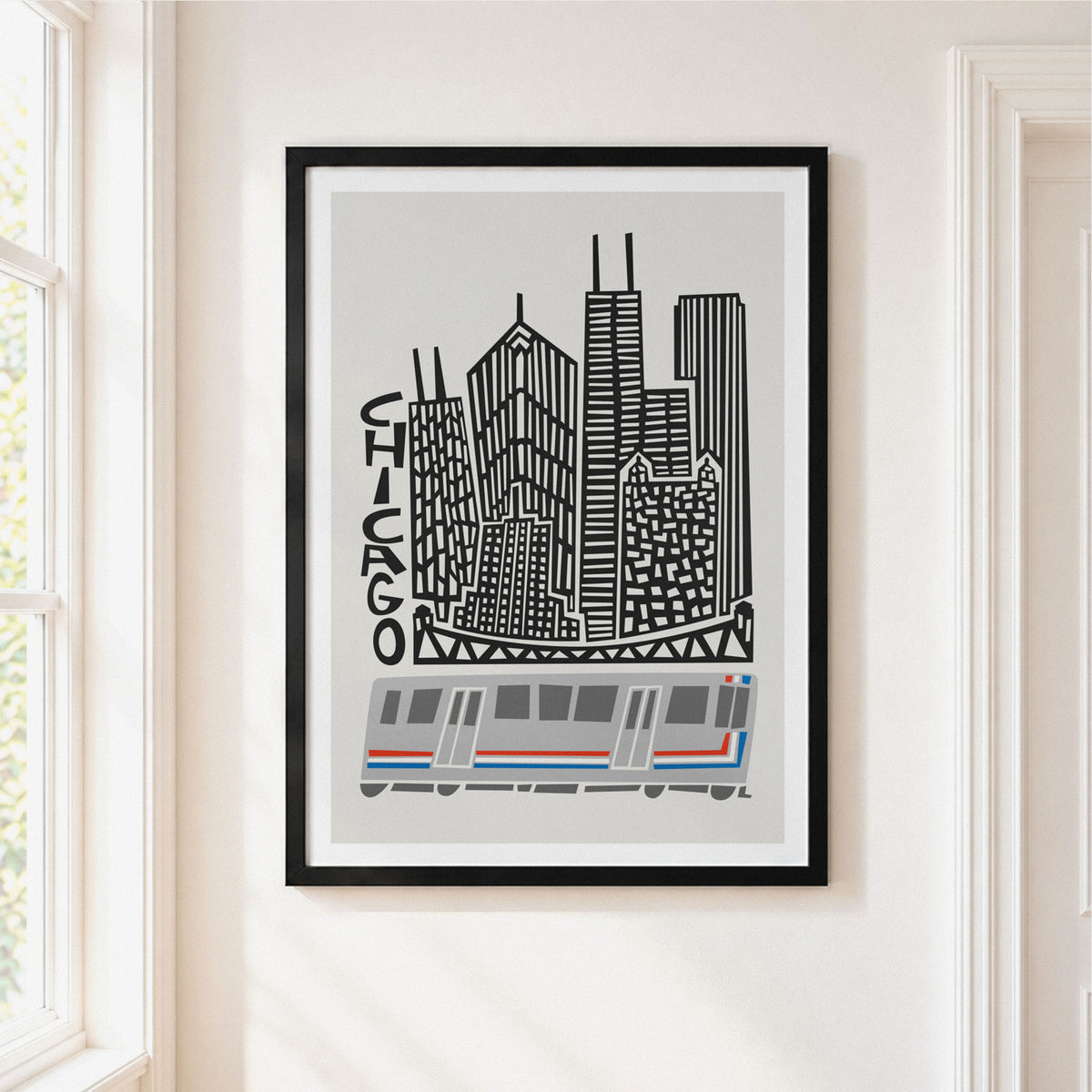 Framed Chicago cityscape wall art hanging on a cream wall beside a window. The print shows black-and-gray skyscrapers with the word CHICAGO and a gray, white, and blue train at the bottom, enclosed in a black frame.