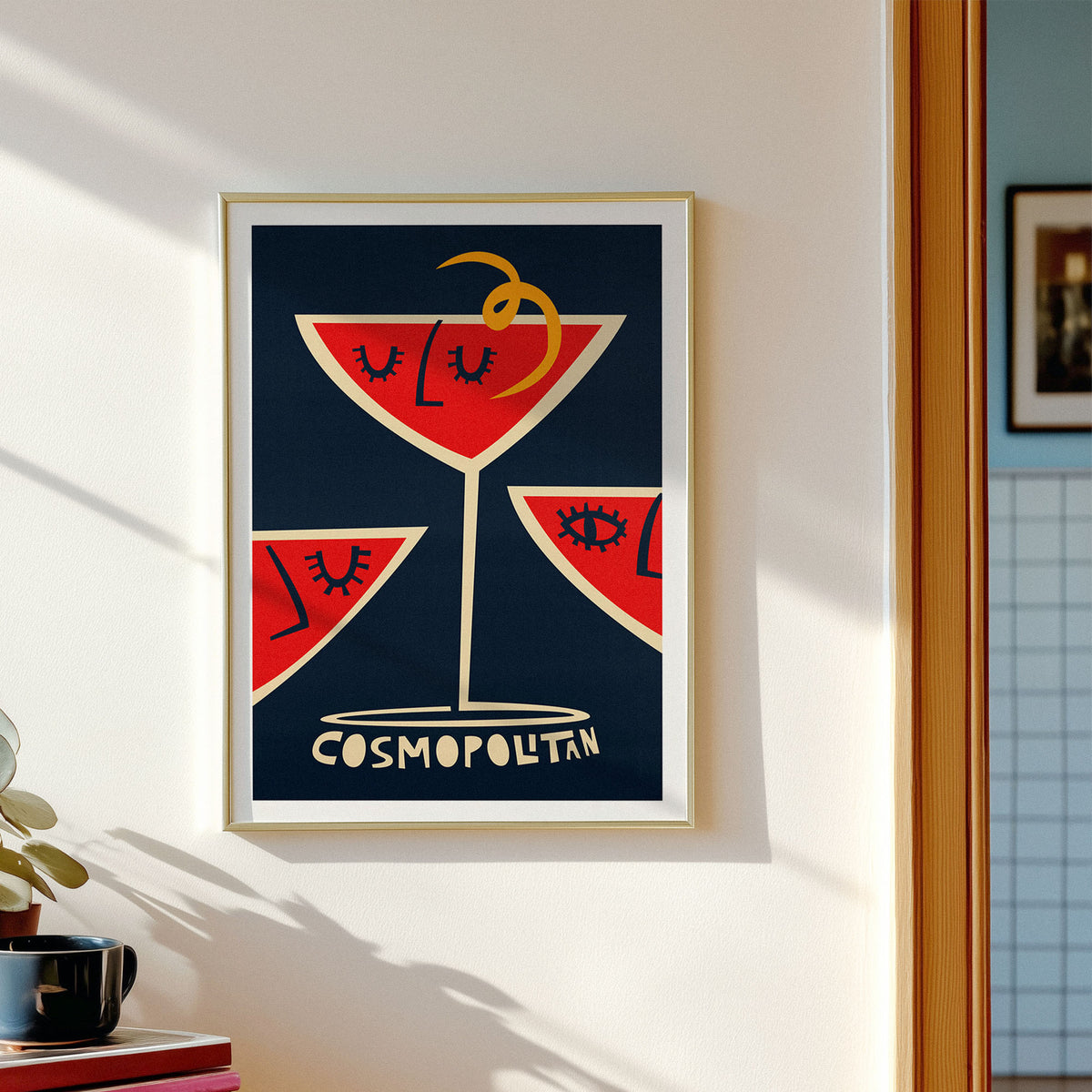 A framed art print featuring a stylized red cocktail glass with closed eyes on a dark navy background, hung on a white wall in a sunlit room. The print includes the word 'COSMOPOLITAN' at the bottom.