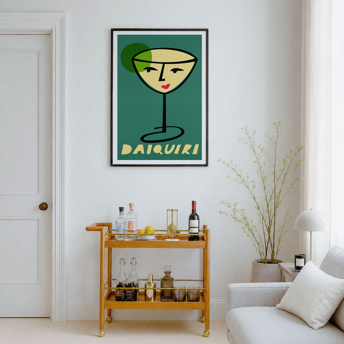 A framed wall art print featuring a stylized cocktail glass with a face and the word “DAIQUIRI” below, displayed in a bright living room above a wooden bar cart and next to a sofa.