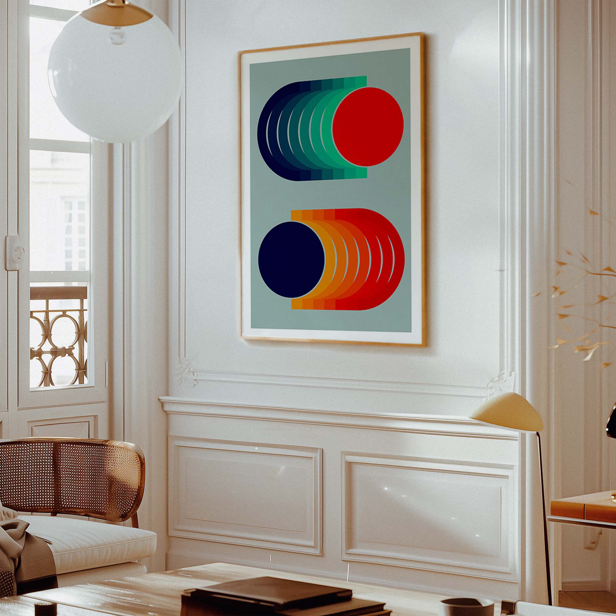 A framed abstract geometric wall art print hangs on a white paneled wall in a bright, modern living room. The artwork features bold, curved shapes in navy, teal, green, red, and orange on a pale blue-gray background, enclosed in a light wood frame.