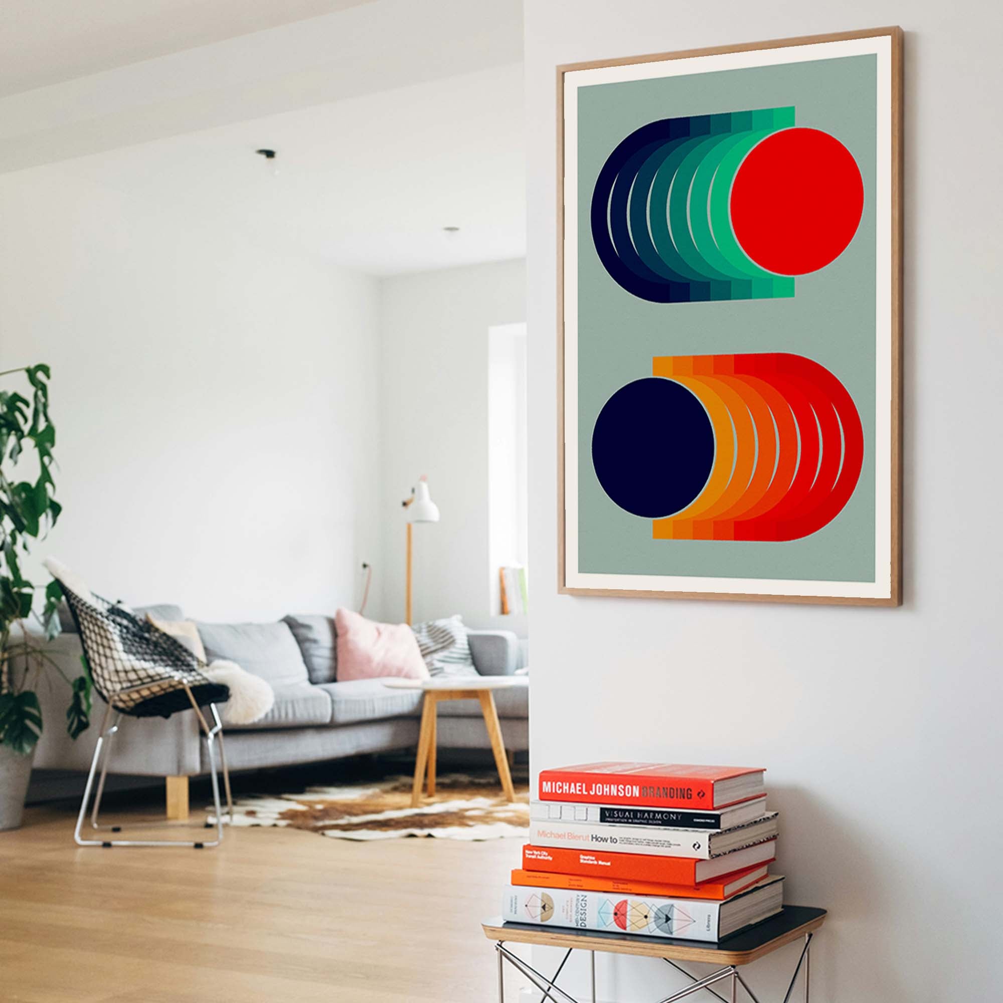Framed abstract wall art hanging on a white wall in a modern living room. The artwork features bold geometric shapes in red, green, navy, orange, and yellow on a muted sage background, enclosed in a light wood frame.