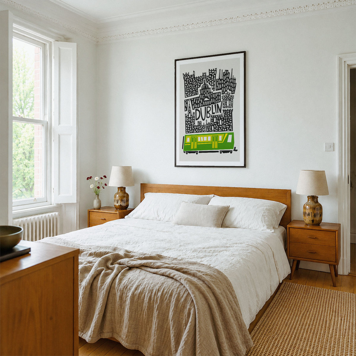 A styled bedroom interior with a wooden bed dressed in white bedding and a beige throw, flanked by matching wooden nightstands and lamps. A framed black-and-white typographic wall print of Dublin with a bright green train hangs above the bed.
