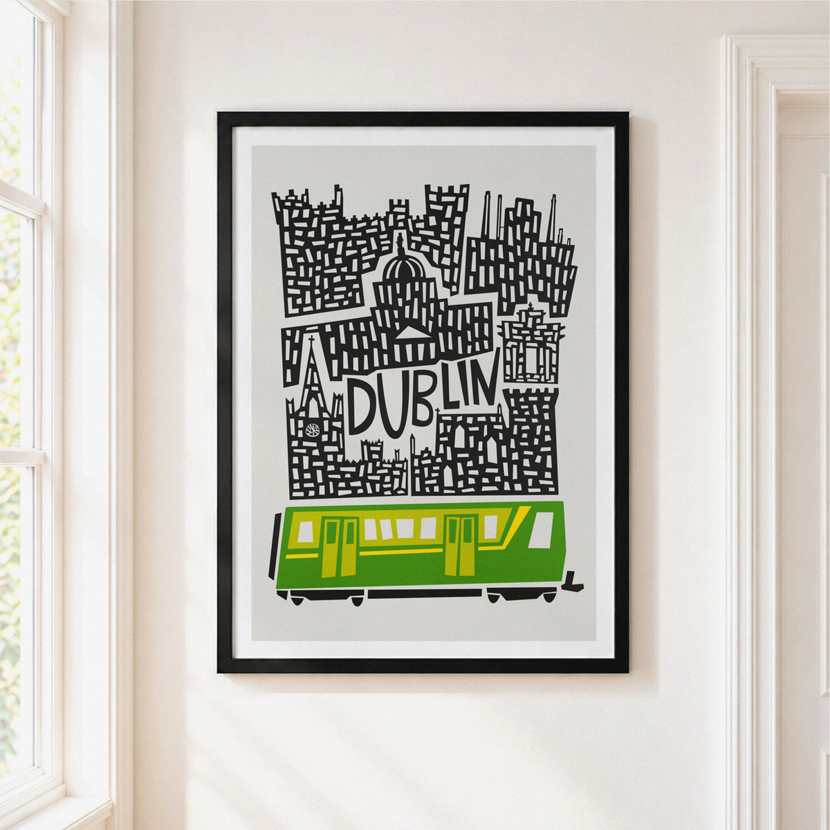 A framed art print featuring a stylized black and white map of Dublin with a green and yellow tram at the bottom, displayed on a white wall in a bright room.
