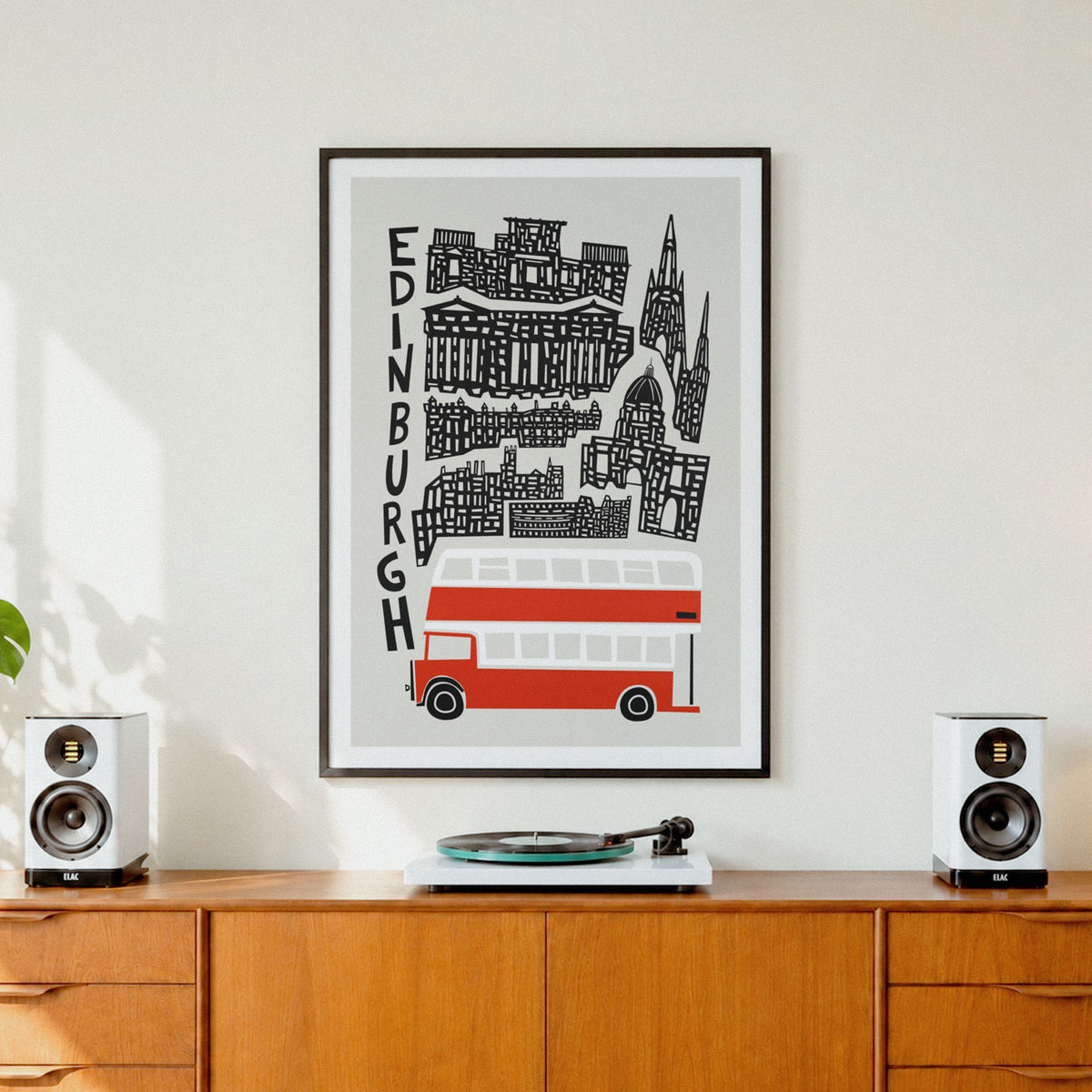 A framed vertical art print featuring a stylized black line drawing of Edinburgh landmarks and a red double-decker bus on a light gray background, displayed on a wooden cabinet between two white speakers.