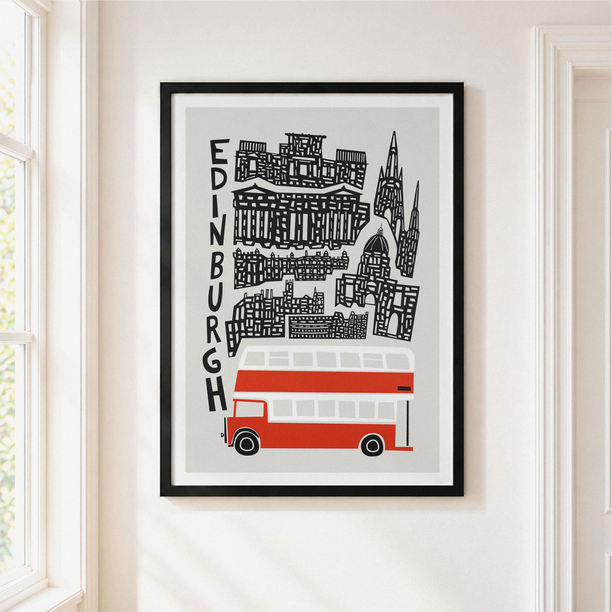 A framed Edinburgh cityscape art print hanging on a white wall beside a window. The poster shows black architectural silhouettes and the word 'EDINBURGH' vertically, with a red and white double-decker bus at the bottom.
