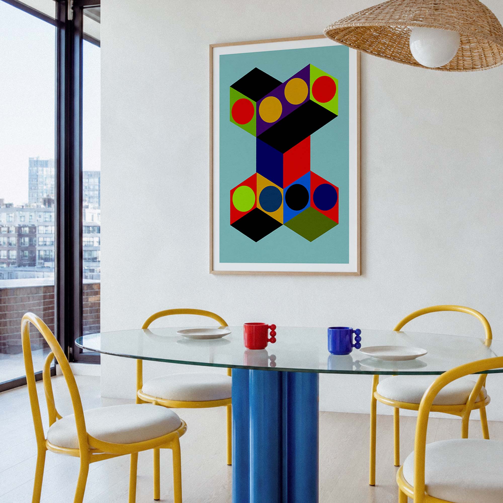 A framed abstract geometric wall art print hangs above a glass dining table with yellow chairs in a bright modern room. The artwork features overlapping black, red, yellow, green, blue, and orange geometric shapes on a light blue background.