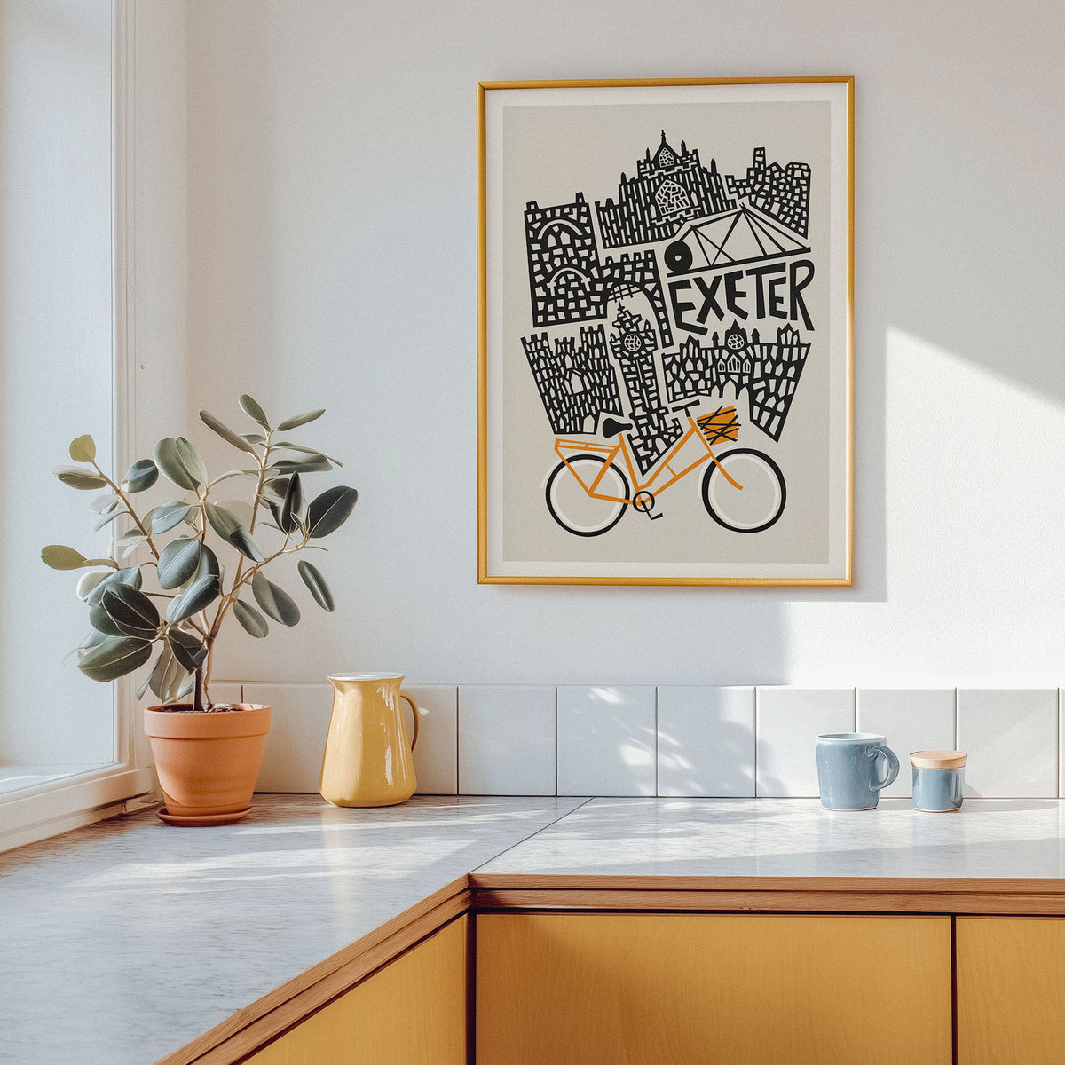 A framed art print featuring a black and white London-style skyline collage with the word 'EXETER' and an orange bicycle, hanging on a white kitchen wall above a countertop with a potted plant and mugs.