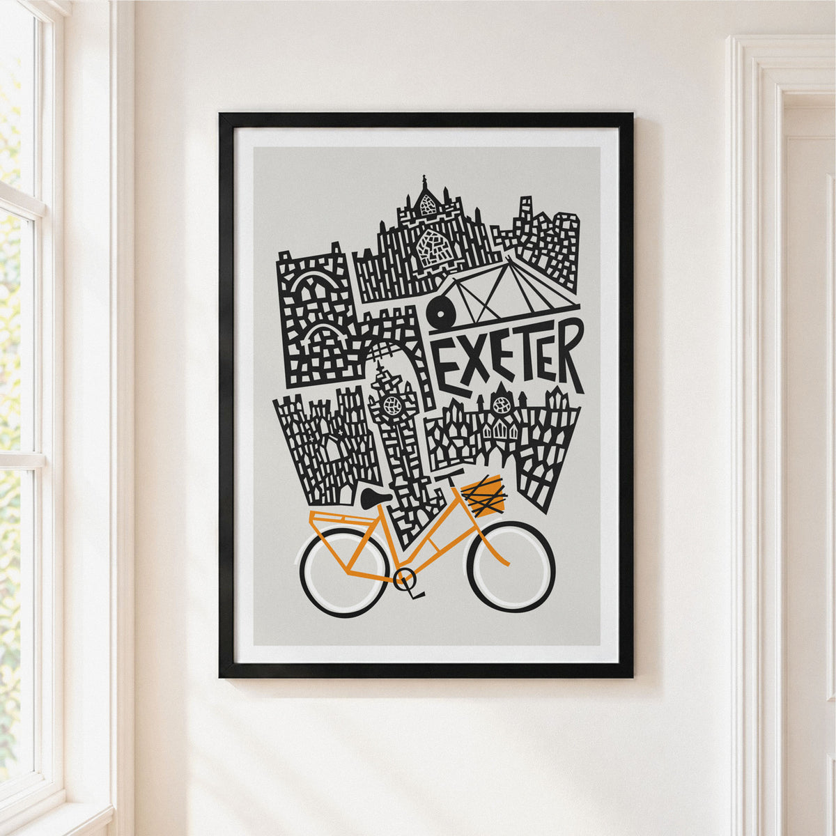 Framed art print featuring a black and white illustrated cityscape with the word 'EXETER' and an orange bicycle, displayed on a light beige wall in a real interior setting.