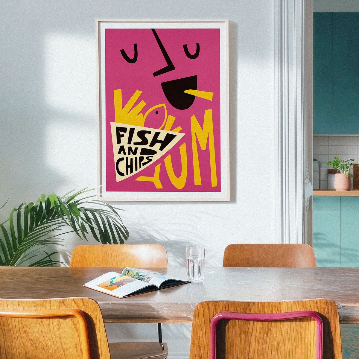 A framed abstract art print featuring a stylized face and the words 'FISH AND CHIPS' in bold yellow and black on a pink background, hanging on a light gray wall above a wooden dining table with chairs in a modern kitchen setting.
