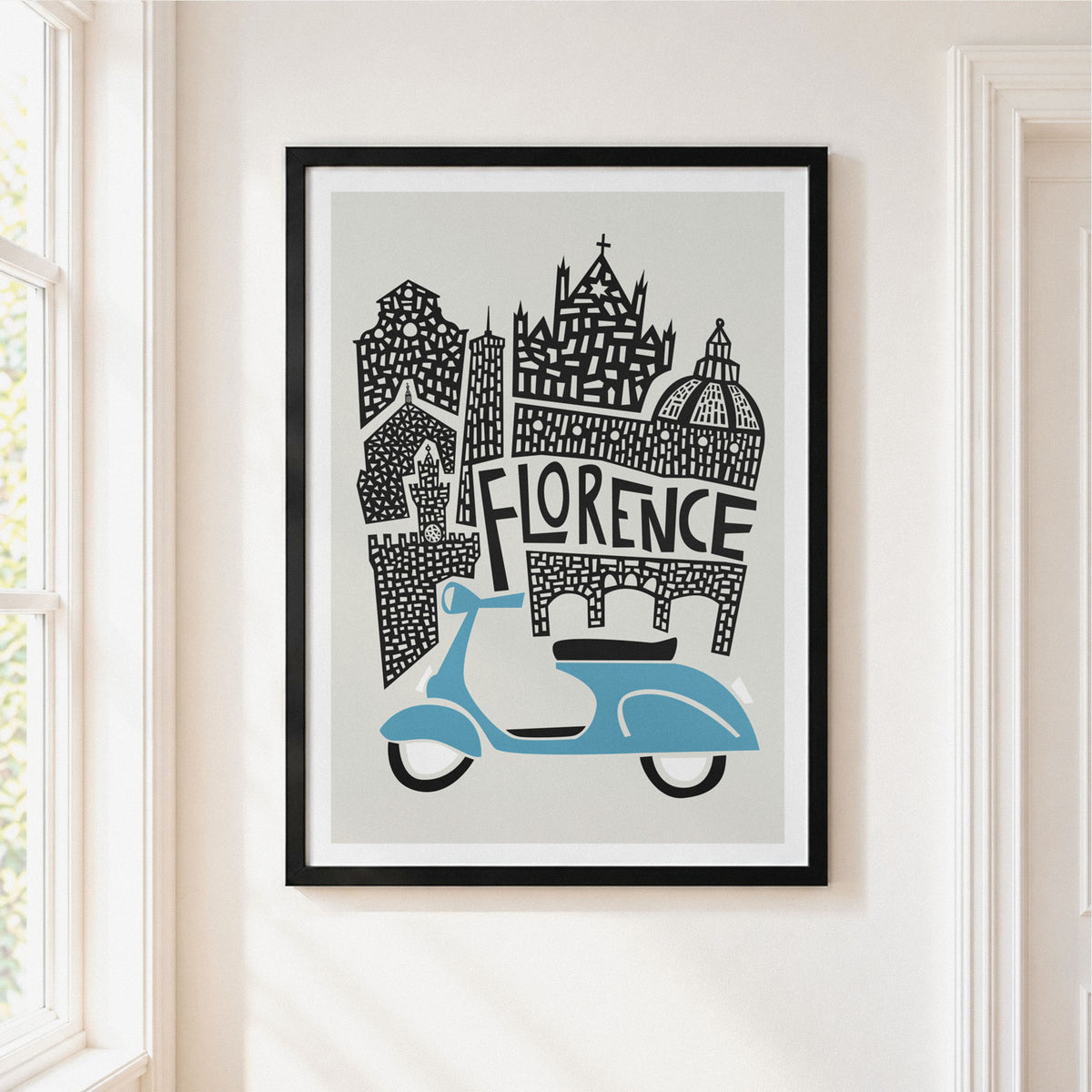A framed art print featuring a stylized black-and-white illustration of Florence architecture with a light blue scooter, hanging on a cream-colored wall next to a window.