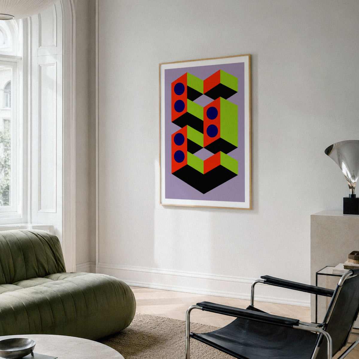 A framed abstract geometric art print with colorful 3D block shapes hangs on a light gray wall in a modern living room setting with a green sofa and black chair.