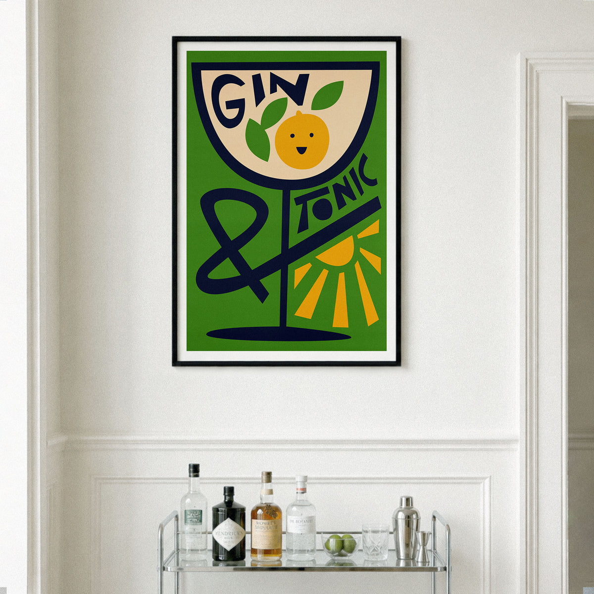 A framed vintage-style wall art print featuring a green background, a tilted glass with the words “GIN TONIC,” and an orange illustration, hanging on a white wall above a small bar cart with bottles and glasses.