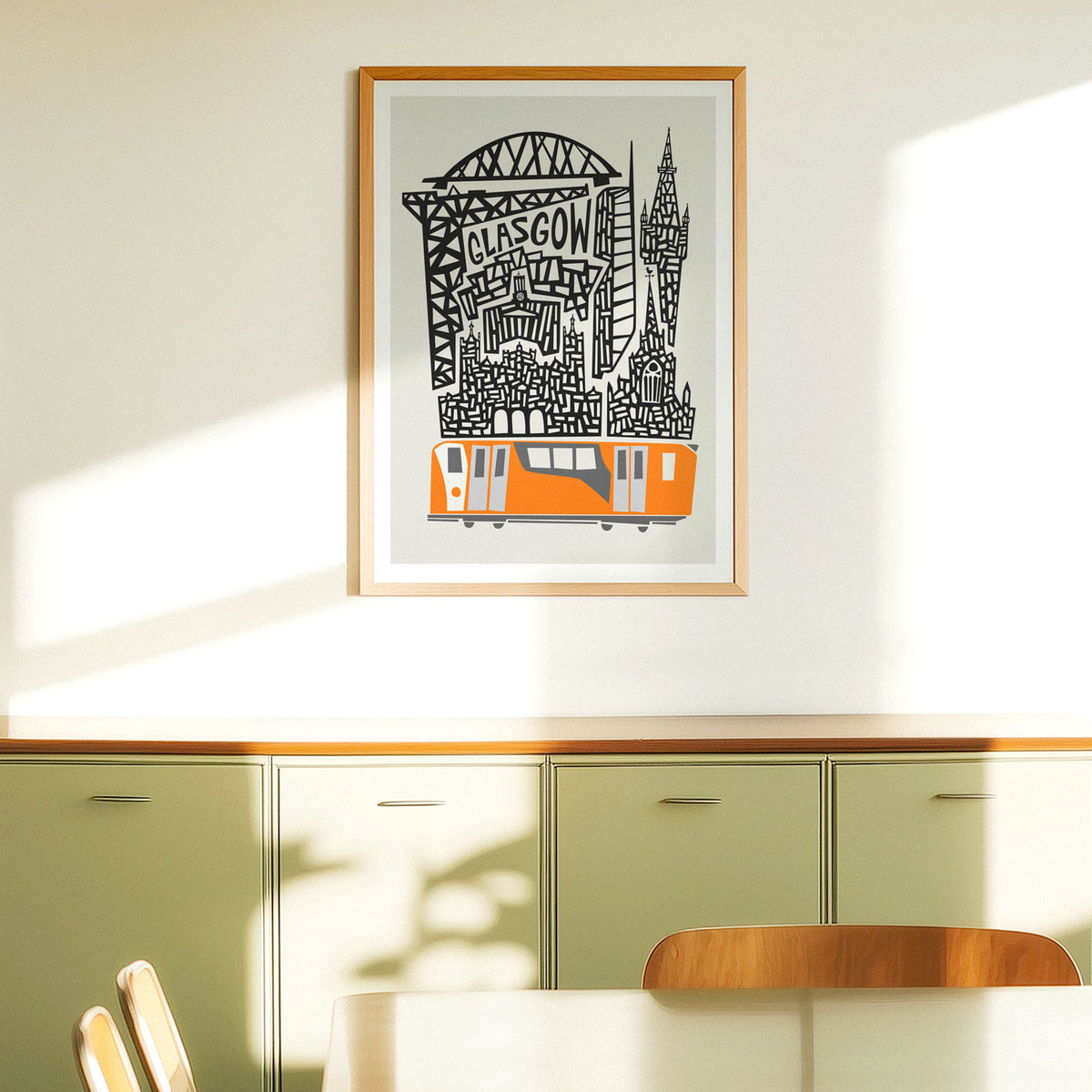 A framed art print featuring a stylized Glasgow skyline with a Orange train and black architectural details on a light gray background, hung on a cream-colored wall next to a window above a sideboard