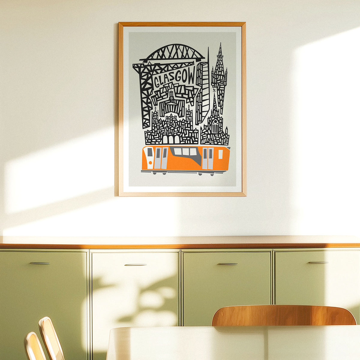 A framed art print featuring a stylized Glasgow skyline with a Orange train and black architectural details on a light gray background, hung on a cream-colored wall next to a window above a sideboard