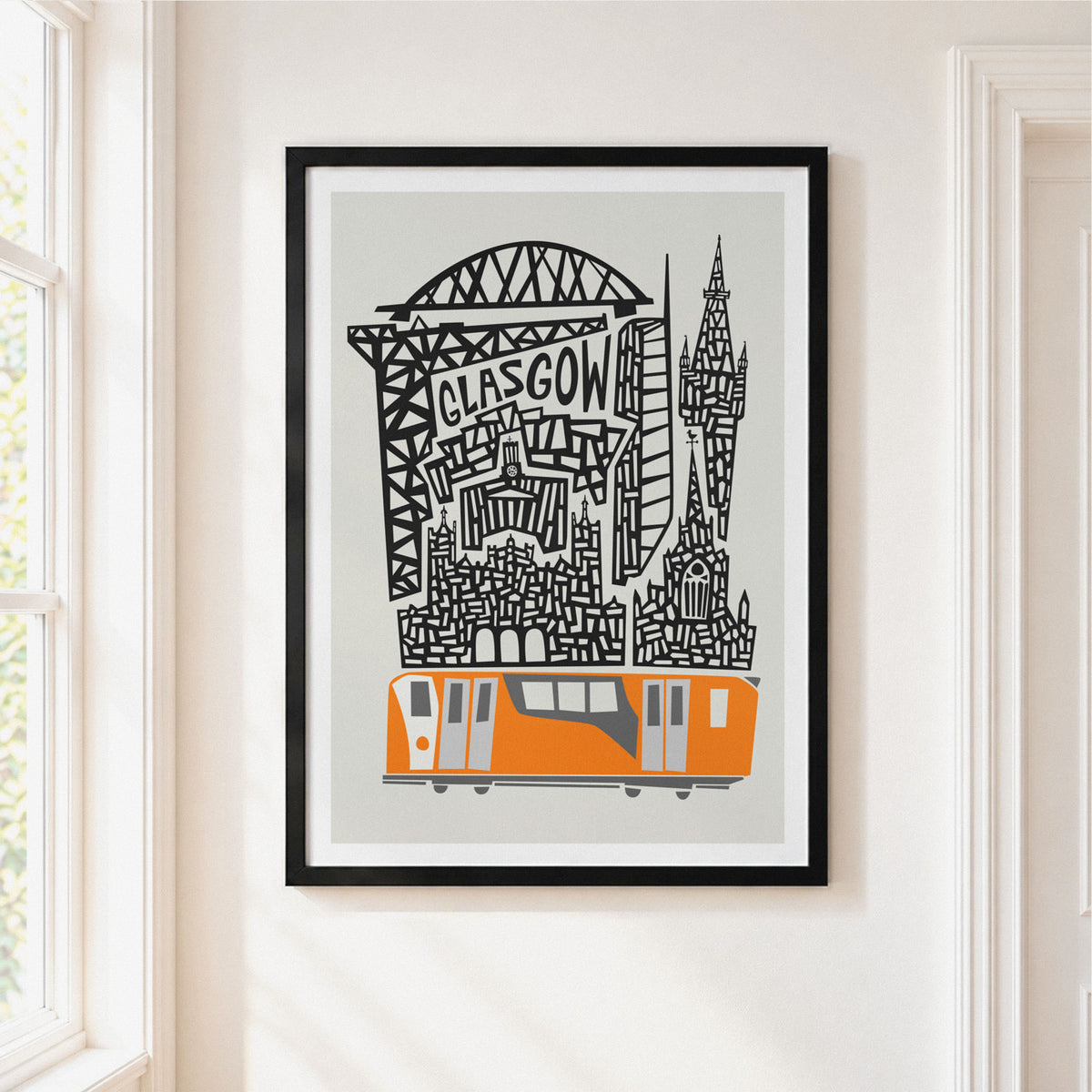 A framed art print featuring a stylized Glasgow skyline with a Orange train and black architectural details on a light gray background, hung on a cream-colored wall next to a window.