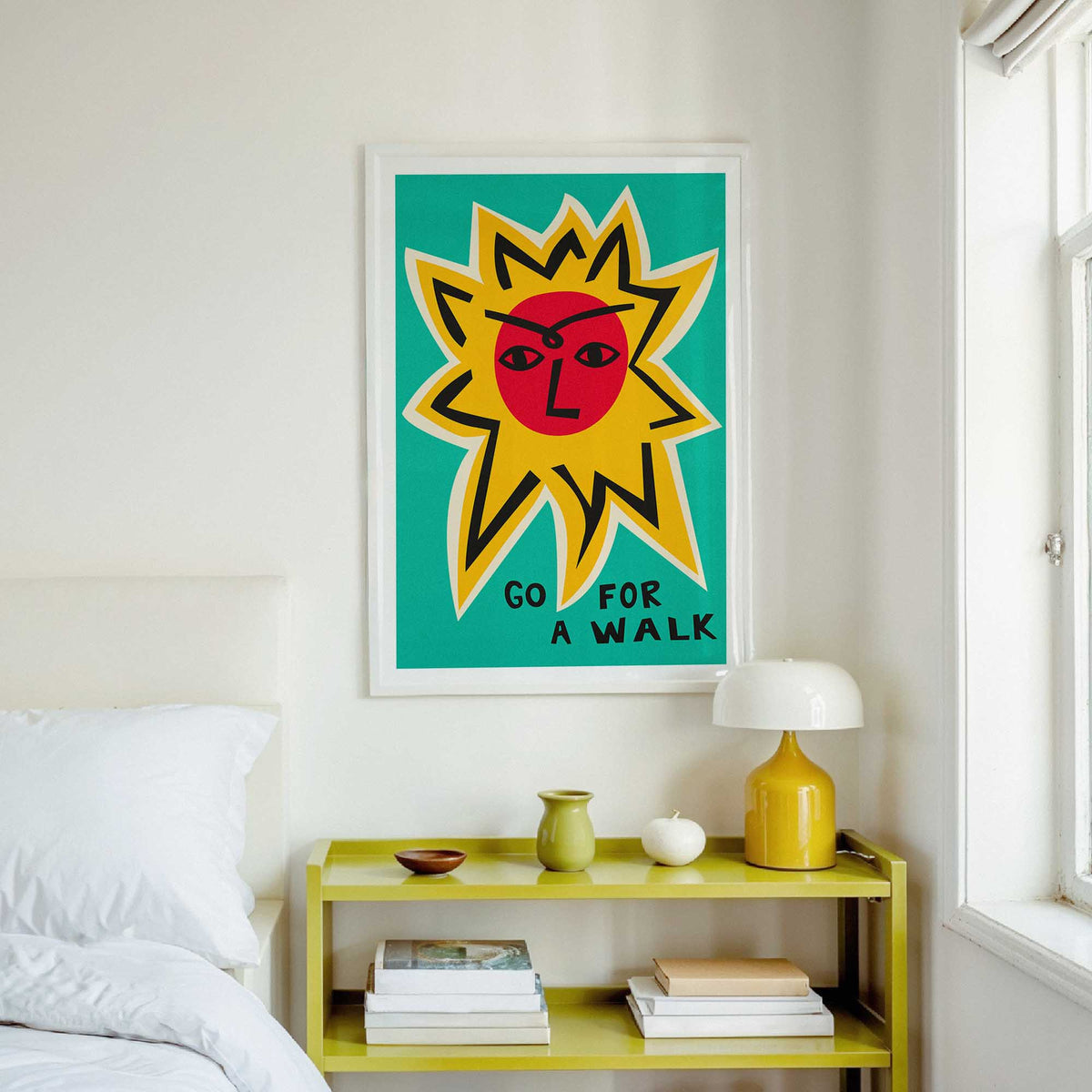 A framed art print featuring a stylized yellow sun with a red face and the typography lettering 'GO FOR A WALK' on a teal background, displayed on a white wall above a yellow bedside table in a bright bedroom.